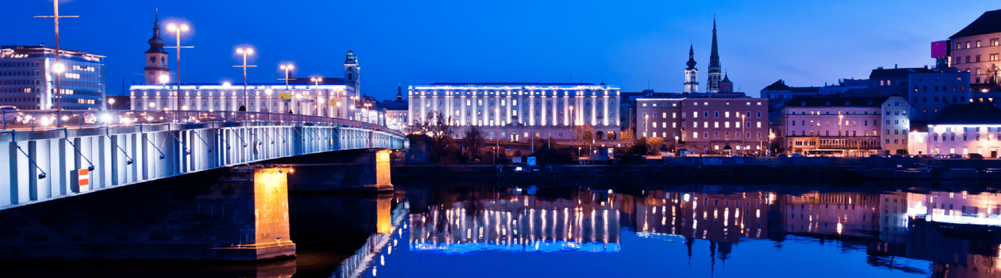 Beleuchtete Donaupromenade in Linz bei Nacht, mit reflektierenden Lichtern auf dem Wasser und stimmungsvoller Atmosphäre.