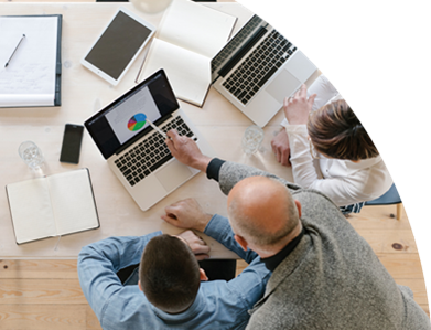 Eine Runde von Personen am Tisch, alle mit Laptops, die gemeinsam arbeiten. Die Person in der Mitte zeigt auf den Laptop auf ein Kreisdiagram.